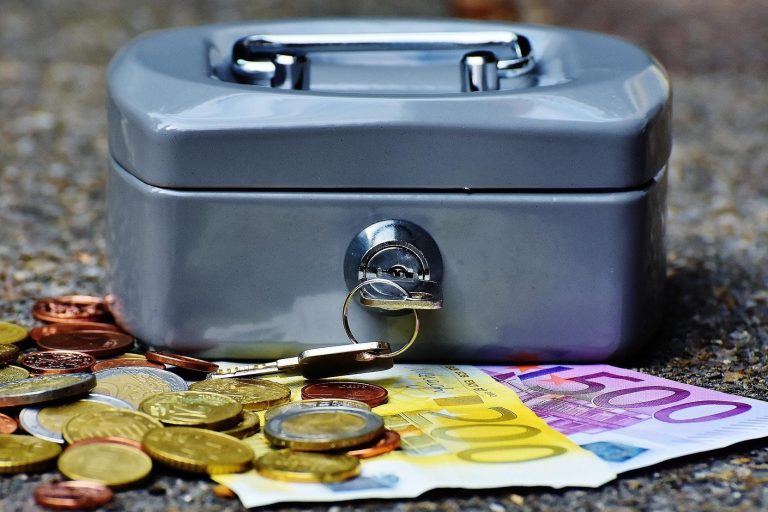 freiwillige Kommunalabgabe Graue Geldkassette mit offenem Schloss und Münzen sowie Euro-Scheinen drumherum.